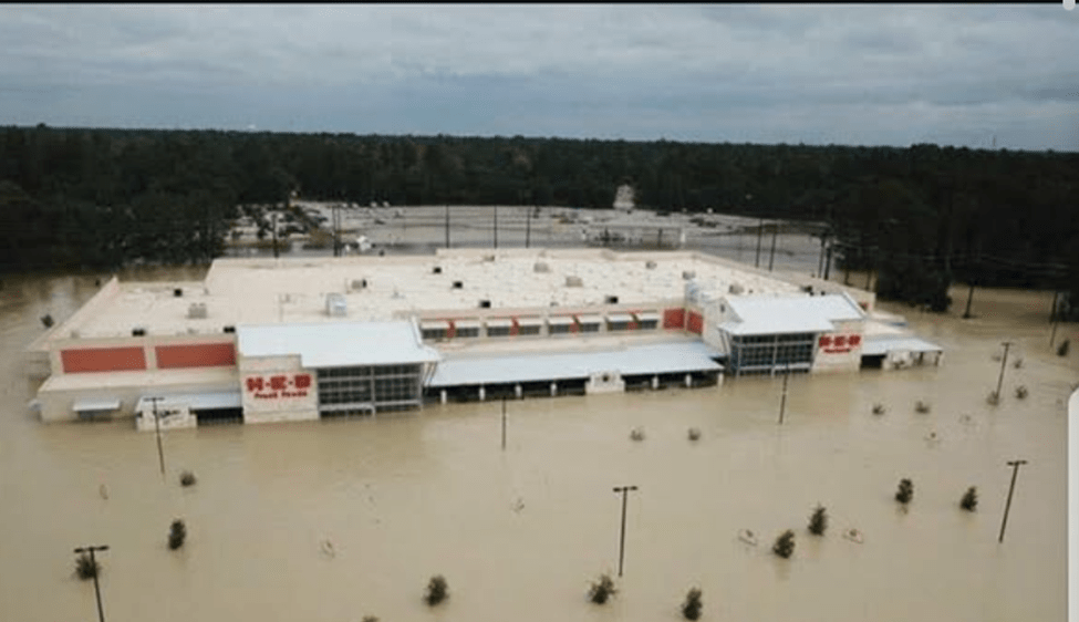 Aerial view of a commercial building partially submerged in floodwater, with surrounding trees and parking lot visible.